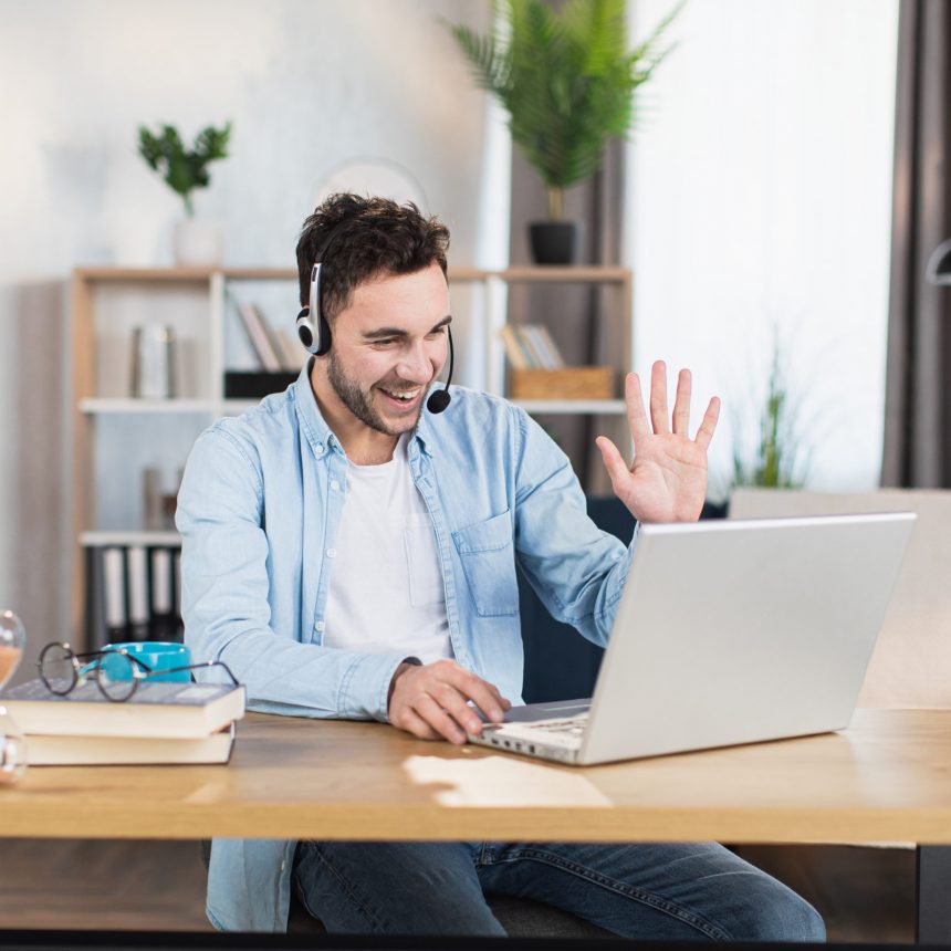 Handsome caucasian man talking and gesturing during video conference on modern laptop. Positive young guy using wireless headset while working at home.